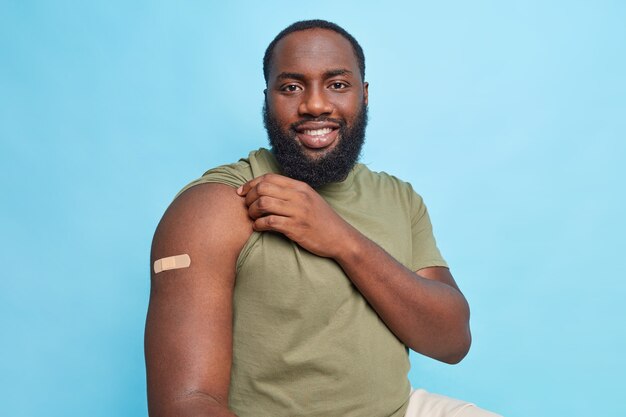 ワクチン接種後の満足した男性は、腕の絆創膏が青い壁で隔離された病気を治すために19回のワクチン接種を受けたことを示しています