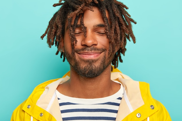 Satisfied man with dark skin, dreads, wears yellow raincoat and striped jumper, keeps eyes closed, imagines something nice