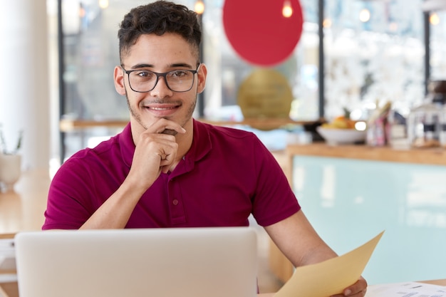 Satisfied man works on clients project, updates software, sits in front of opened laptop computer, holds document, keeps hand on chin, models over cozy cafeteria interior, thinks on idea for startup