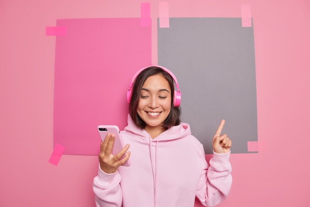 Satisfied millennial Asian woman makes video call promots something indicates at blank space smiles pleasantly shows direction sale logo store banner wears hoodie poses against pink wall