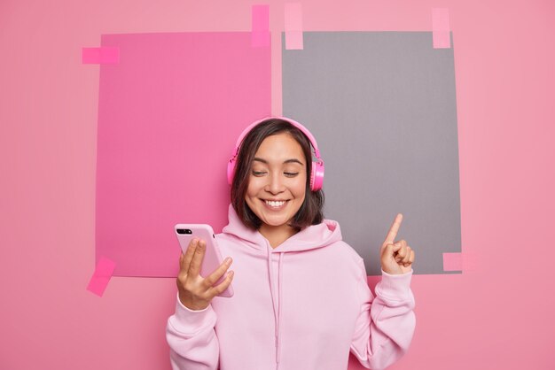 만족스러운 밀레니엄 아시아 여성은 빈 공간에서 무언가를 나타내는 화상 통화를 하고 미소는 유쾌하게 방향 판매 로고 매장 배너가 분홍색 벽에 후드티 포즈를 취하는 것을 보여줍니다