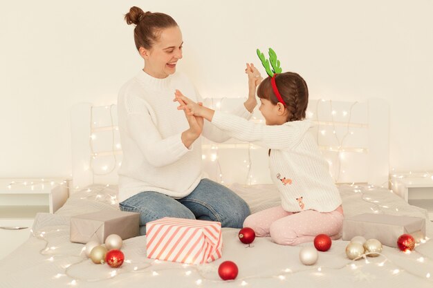 Довольные мать и дочь в белых свитерах повседневного стиля сидят на кровати, держась за руки, празднуют зимние праздники, семья с праздничным настроением празднует новый год.