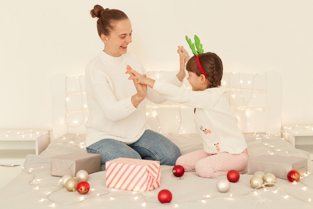 Satisfied mother and daughter wearing casual style white sweaters sitting on bed holding each other hands, celebrating winter holidays, family with festive mood celebrating new year.
