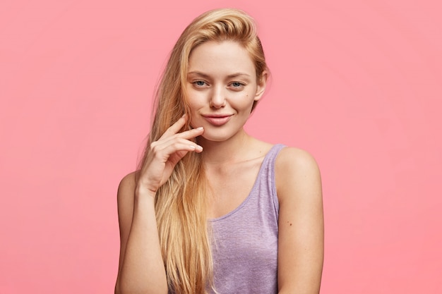 Satisfied mysterious blonde blue eyed young woman has delighted expression, has luxurious light hair, wears casual purple t shirt, thinks about something pleasant