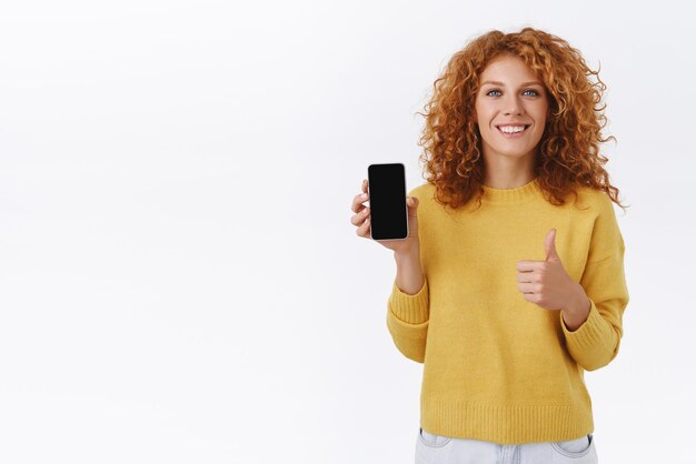 Satisfied pleased caucasian ginger curly girl in yellow sweater holding smartphone promote good mobile application show thumbup and smiling nod in approval advice download app