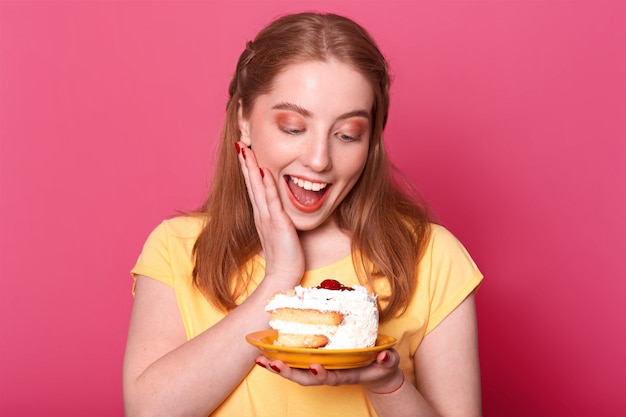 довольная довольная девушка со светло-каштановыми волосами, держит огромный кусок вкусного торта, держит рот открытым, полный удовольствия, одетый в повседневную желтую футболку