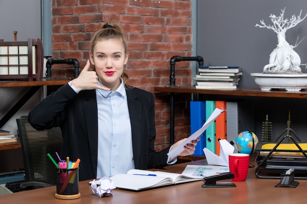 Satisfied and positive young lady sitting at a table and holding the document making call me gesture in the office