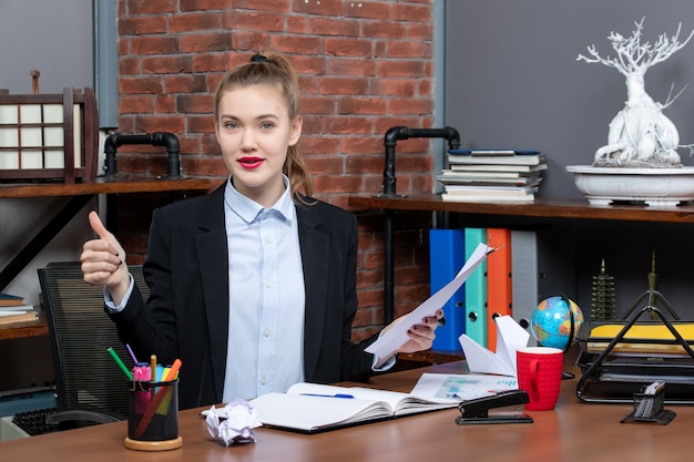 Free photo satisfied and positive young lady sitting at a table and holding the document making ok gesture in the office