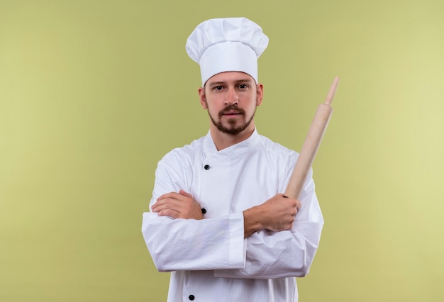 満足しているプロの男性シェフが白い制服を着て調理し、腕を組んで立っている緑の背景に自信を持って麺棒を保持している帽子を調理