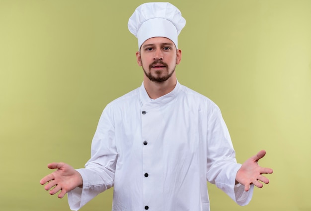 Satisfied professional male chef cook in white uniform and cook hat spreading hands making welcoming gesture standing over green background