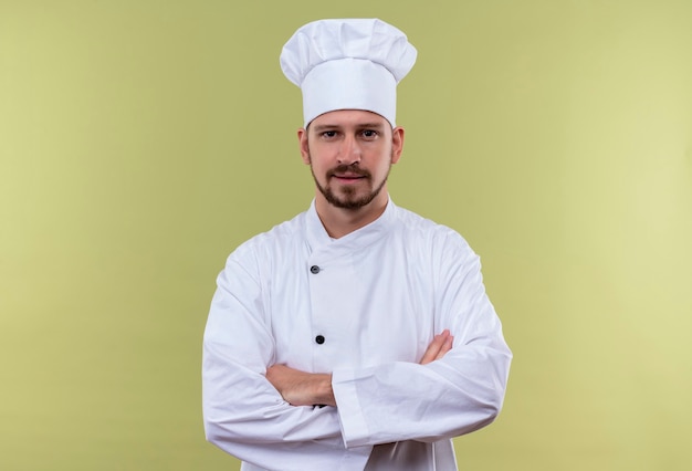 Удовлетворенный профессиональный шеф-повар-мужчина в белой форме и поварской шляпе, стоя со скрещенными руками, уверенно глядя на зеленом фоне