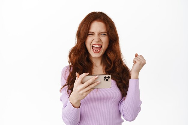 Satisfied redhead woman celebrating victory, say yeah yes and fist pump, winning on mobile phone, hold smartphone horizontally, standing against white background