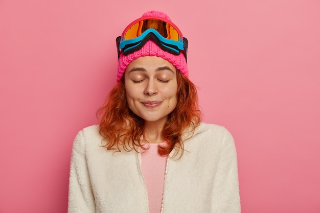 Free photo satisfied redhead woman wears warm hat, ski goggles and white jumper