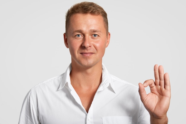 Satisfied self assured handsome man with confident expression, makes okay gesture, shows his agreement with something, dressed in elegant white shirt, poses in studio. Body language concept.