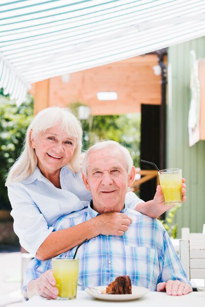 Satisfied senior couple hugging in cafe on terrace enjoying refreshing drink and cake