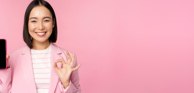 Free photo satisfied smiling asian businesswoman recommending mobile phone app website company on smartphone showing screen and okay sign pink background