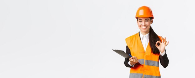 Удовлетворенная улыбающаяся азиатская женщина-архитектор-строитель в шлеме и светоотражающей куртке, показывающая хороший жест, одобряет и дает разрешение на завершение строительных работ