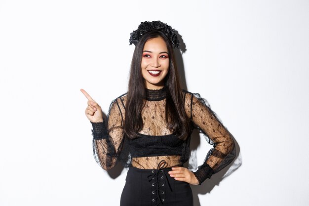 Satisfied smiling asian woman in evil witch or banshee costume celebrating halloween, looking pleased and pointing finger upper left corner, showing your promo banner, white background.