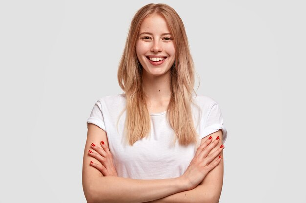 Довольно улыбающаяся молодая блондинка с довольным выражением лица выражает счастье, одетая в повседневную белую футболку