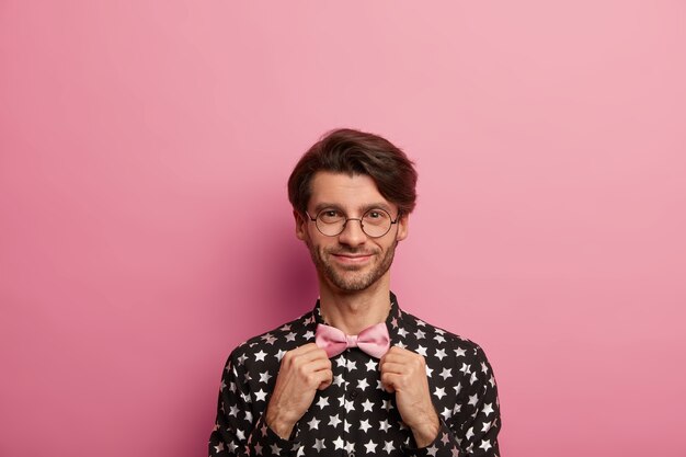 Satisfied unshaven fashionable man holds hands on pink bowtie, looks gladfully at himself, dresses for date or meeting, wears optical round specs