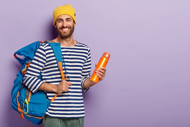 Free photo satisfied unshaven man holds thermos and big rucksack, ready for adventurous trip, smiles gladfully, dressed in casual wear, enjoys hot drink isolated over purple background free space for your advert