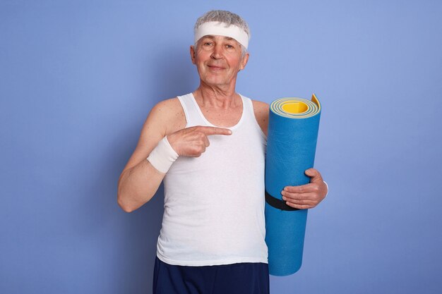 Satisfied white haired male with yoga mat posing isolated, pointing with index finger aside, wearing sleeveless t-shirt, hairband and wristband.
