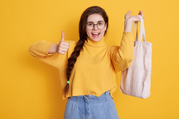 Satisfied woman with long pigtail showing thumb up and holding bag in hand, enjoying her shopping