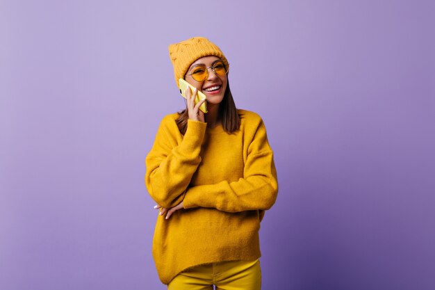 Satisfied woman with shoulder long hair happy while talking with best friend by smartphone. Genuinely smiling student in stylish hat and yellow outfit wants to make snapportrait on isolated purple