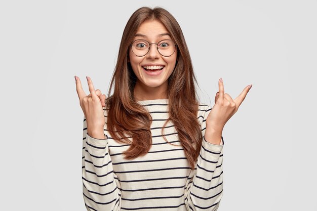 Satisfied woman with toothy smile, raises both hands and shows rock n roll sign, dressed in striped jumper, models against white wall, listens heavy music, likes rock. Hipster enjoys music