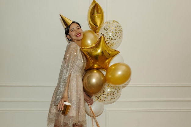 Free photo satisfied young asian girl in beige dress holds bunch of golden balloons on white background holiday concept