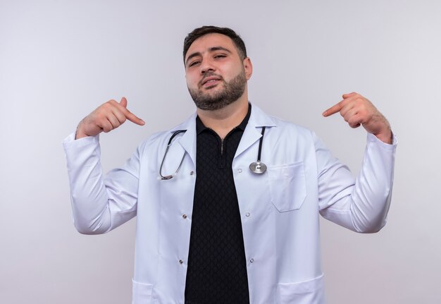 Satisfied young bearded male doctor wearing white coat with stethoscope pointing to himself looking confident 