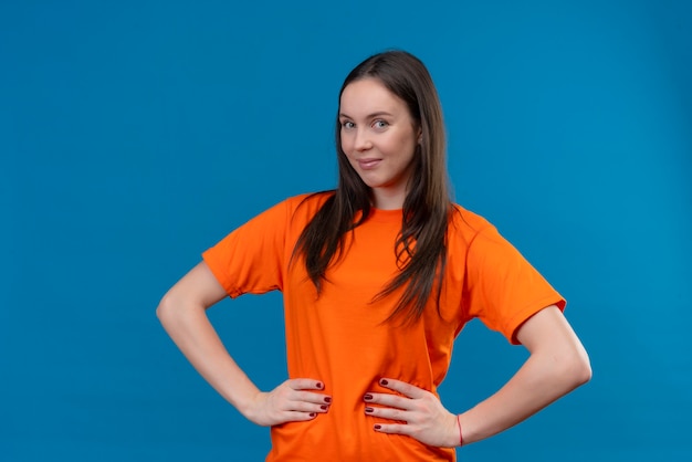 Foto gratuita giovane bella ragazza soddisfatta che indossa la maglietta arancione che guarda l'obbiettivo con un sorriso fiducioso sul viso in piedi su sfondo blu isolato