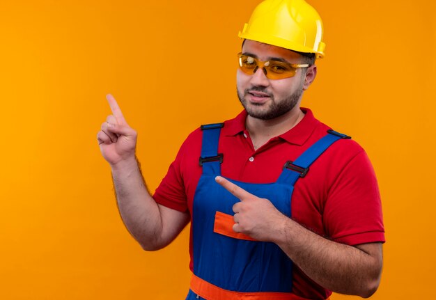Satisfied young builder man in construction uniform and safety helmet looking confident pointing with fingers to the side 