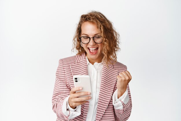 Satisfied young business woman looking at mobile phone and celebrating, triumphing, making money on smartphone, standing white