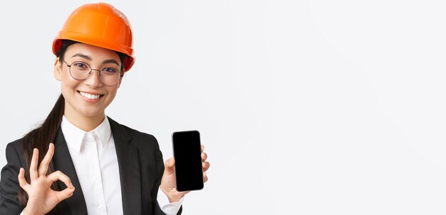 Satisfied young female asian engineer architect in suit and safety helmet showing mobile phone screen and make okay gesture approve recommend application white background