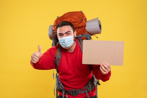 Удовлетворенный молодой парень в медицинской маске с рюкзаком и держит простыню без письма, делая жест на изолированном желтом фоне