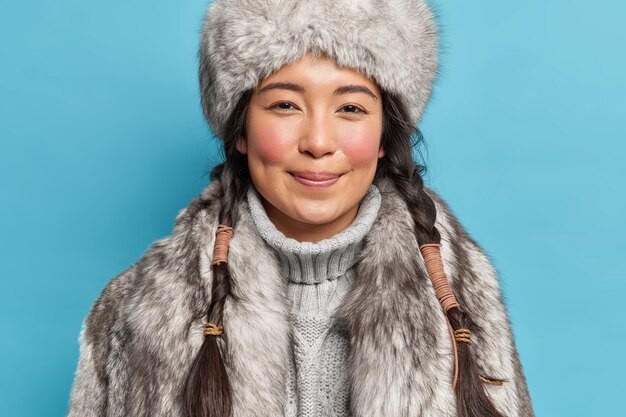 satisfied young siberia woman with two pigtails rosy cheeks smiles pleasantly at front dresses for cold polar weather conditions isolated over blue wall