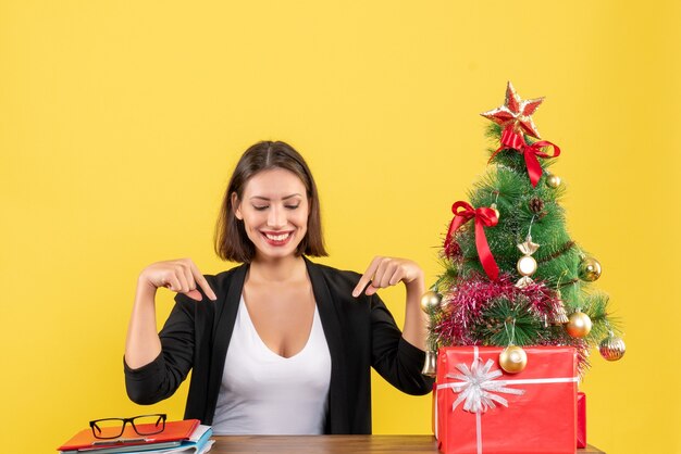 노란색 사무실에서 장식 된 크리스마스 트리 근처 소송에서 아래쪽을 가리키는 만족 된 젊은 여자