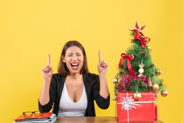 Satisfied young woman pointing up in suit near decorated Christmas tree at office on yellow 