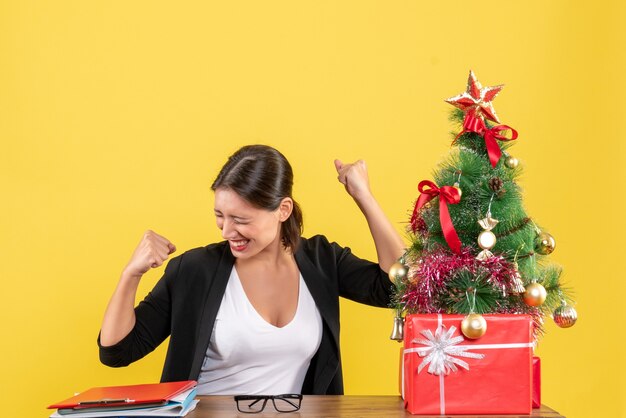 Satisfied young woman in suit near decorated Christmas tree at office on yellow 