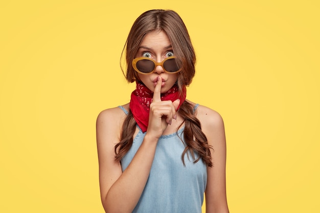 Free photo do not say this information to anyone. attractive woman has two plaits, makes shush gesture, demands keep quiet and be silent, wears sunglasses, fashionable outfit, models indoor over yellow wall