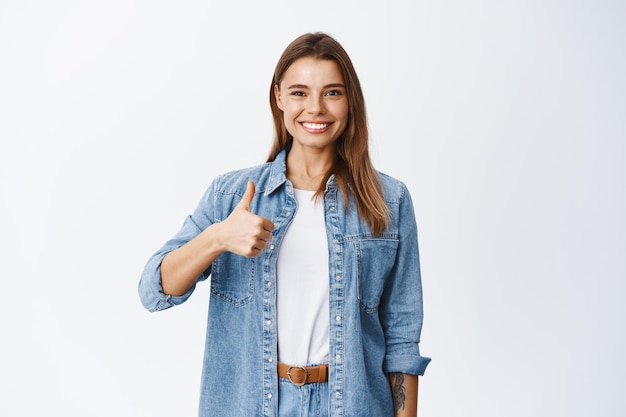 Foto gratuita dire di sì. sorridente donna positiva con capelli biondi e vestiti in denim, che mostra il pollice per apprezzare e approvare, lodare le cose buone, consigliare il prodotto, bianco