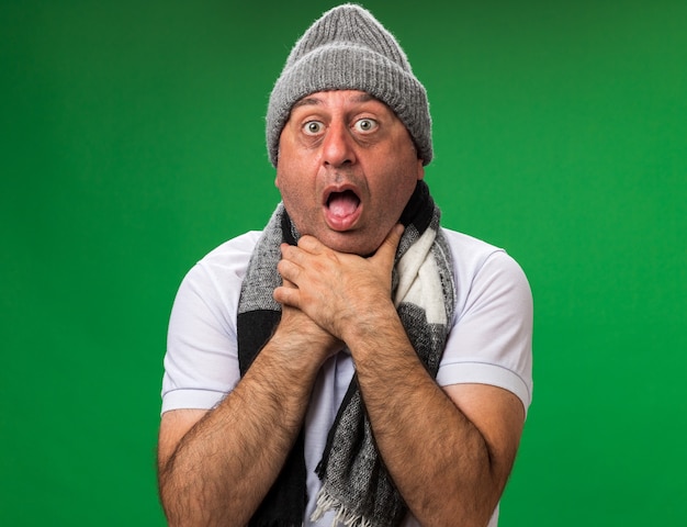 Бесплатное фото Испуганный взрослый больной кавказский мужчина с шарфом на шее в зимней шапке душит себя руками, изолированными на зеленой стене с копией пространства