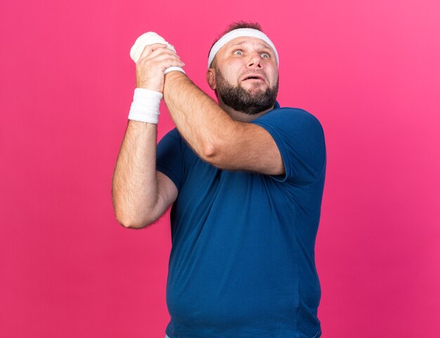 Free photo scared adult slavic sporty man wearing headband and wristbands holding his hand and looking up isolated on pink wall with copy space