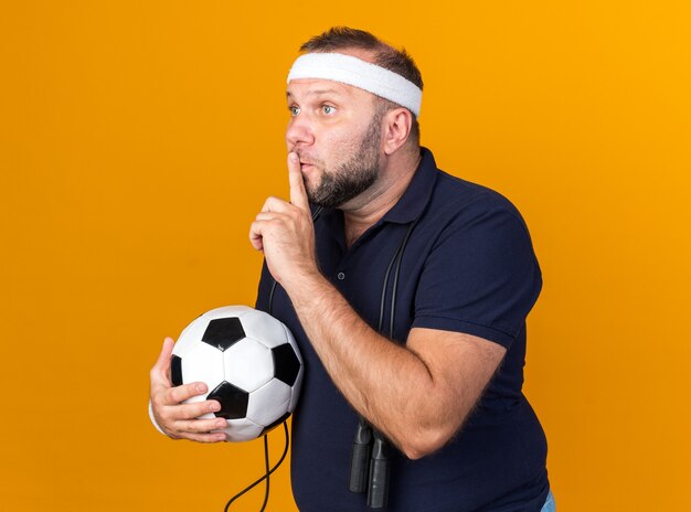 scared adult slavic sporty man with jumping rope around neck wearing headband and wristbands holding ball and doing silence gesture isolated on orange wall with copy space