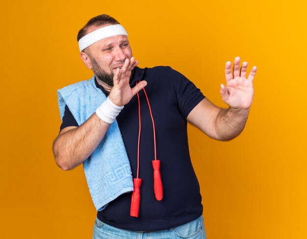 Scared adult slavic sporty man with jumping rope around neck wearing headband and wristbands holding towel on shoulder and keeping hands open looking at side on orange wall with copy space