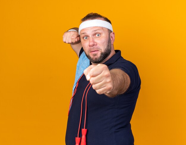 испуганный взрослый славянский спортивный мужчина со скакалкой на шее и с полотенцем на плече, с повязкой на голову и браслетами, держащими кулаки в готовности к удару, изолирован на оранжевой стене с копией пространства