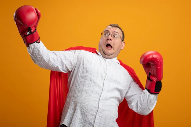 Scared adult slavic superhero man in red cape wearing glasses and box gloves looking at side keeping hands in air isolated on orange background