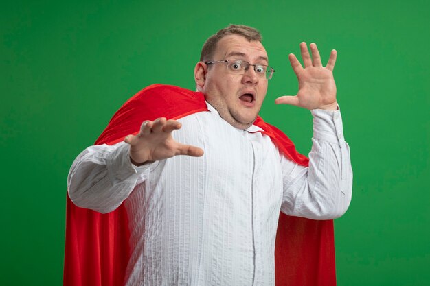 Испуганный взрослый славянский супергерой в красной накидке в очках, держа руки в воздухе, глядя в камеру, изолированную на зеленом фоне