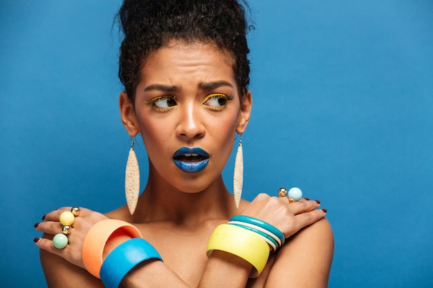 Free photo scared or agitated mulatto woman with colorful makeup and fashion accessories looking aside with crossed hands on shoulders, over blue wall